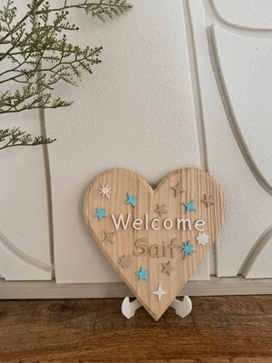 Wooden heart-shaped sign with 'Welcome Saif' text on a stand against a white wall.
