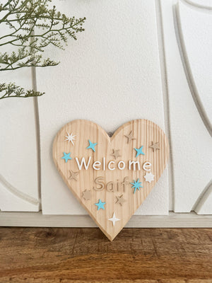 Wooden heart-shaped sign with 'Welcome Saif' text and star decorations on a wooden surface.