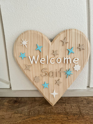 Wooden heart-shaped sign with 'Welcome Saif' text and decorative stars on a white wall.