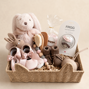 A baby gift basket arranged in a woven natural fiber basket, featuring a plush bunny, soft pink baby shoes with bows, a wooden bead brush, a small brown knit item, a packaged pacifier clip, and a brown and blush pink FRIGG pacifiers accompanied by two candy bags.