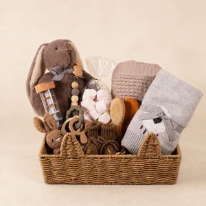 A baby gift basket filled with soft baby items, including a plush bunny toy, a gray knitted wool blanket, a folded gray blanket or swaddle, wooden teething rings, a small wooden bottle, and a pink plush toy, arranged in a woven wicker basket against a warm beige background.
