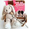 Baby gift set with plush toy, shoes, and 'Hello My Name is Hind' sign in a basket on a white background.