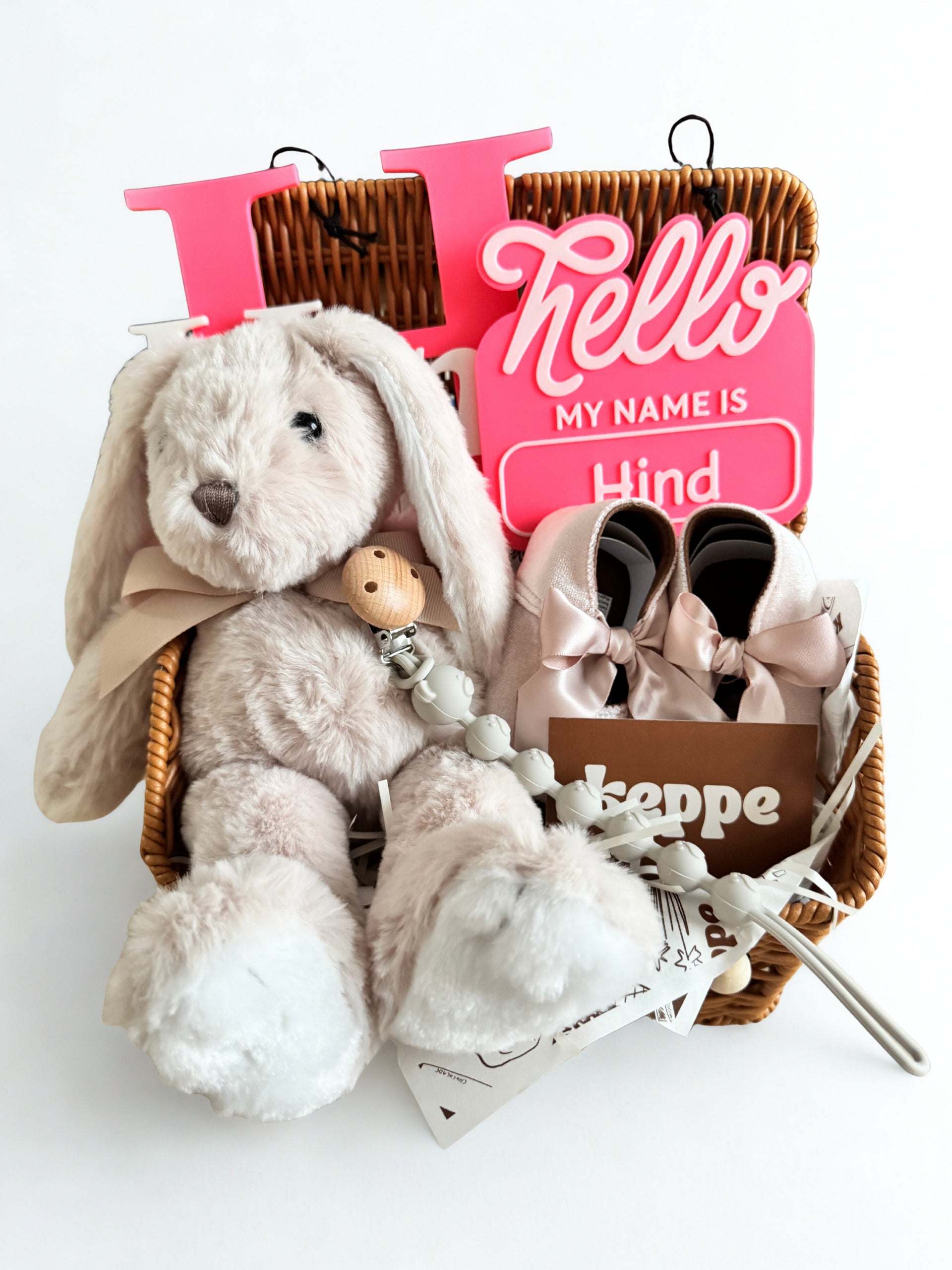 Baby gift set with plush toy, shoes, and 'Hello My Name is Hind' sign in a basket on a white background.